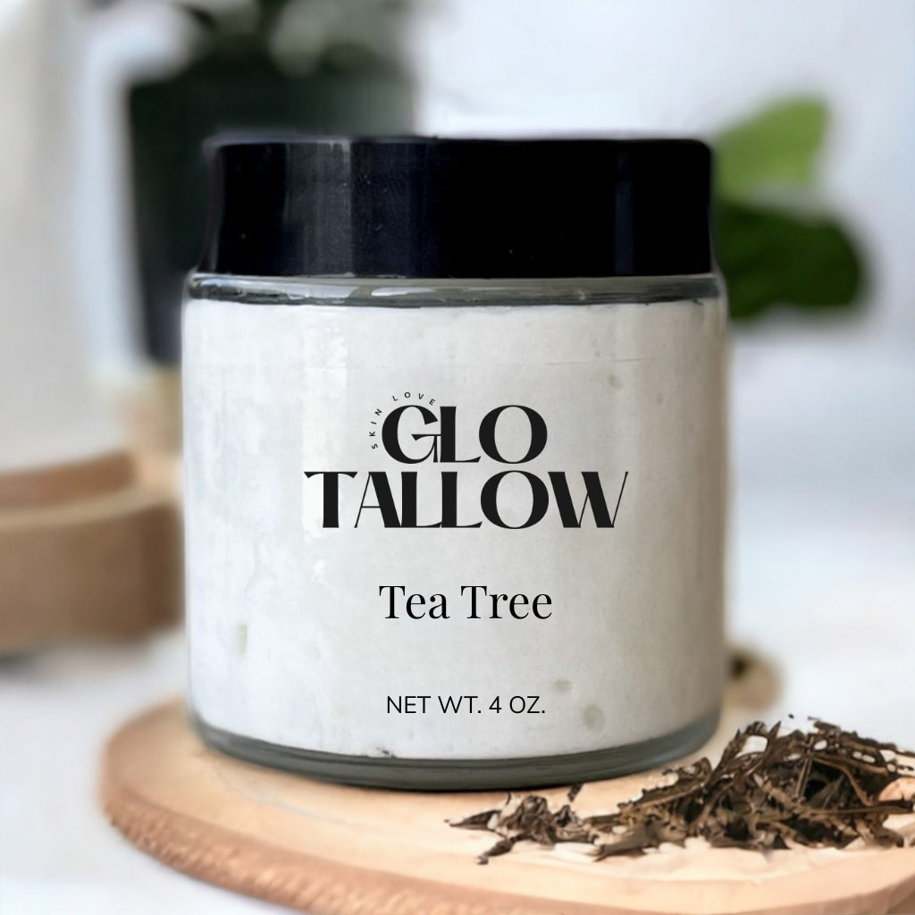 A jar of Glo Tallow Tea Tree balm placed on a wooden surface with a label that includes the product name and weight, net 4 oz. Some tea tree leaves are scattered in front of the jar.