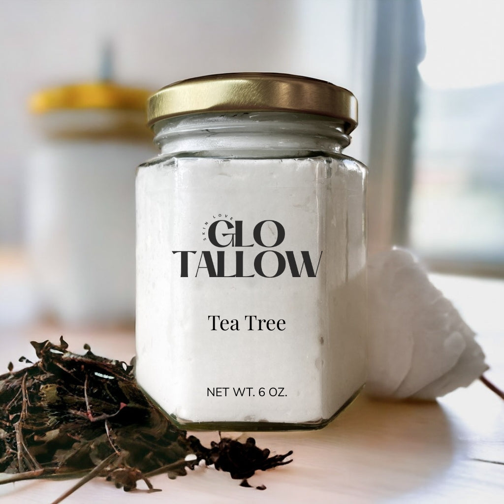 A jar of Glo Tallow Tea Tree balm placed on a wooden surface with a label that includes the product name and weight, net 6 oz. Some tea tree leaves are scattered in front of the jar.